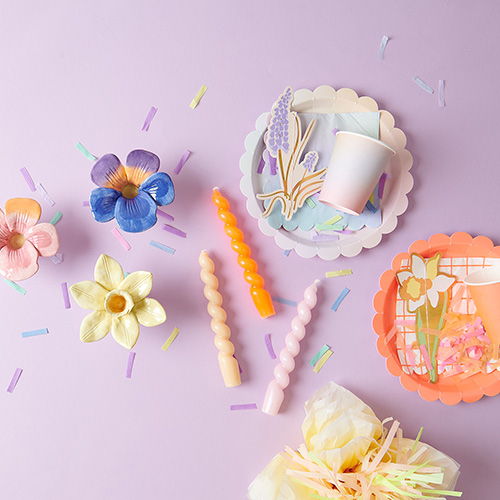 Pappteller darauf Servietten, Becher und Papierblumen, daneben Stabkerzen mit passenden Kerzenhaltern in Blütenform. Produkte aus der Kollektion Futschikato.