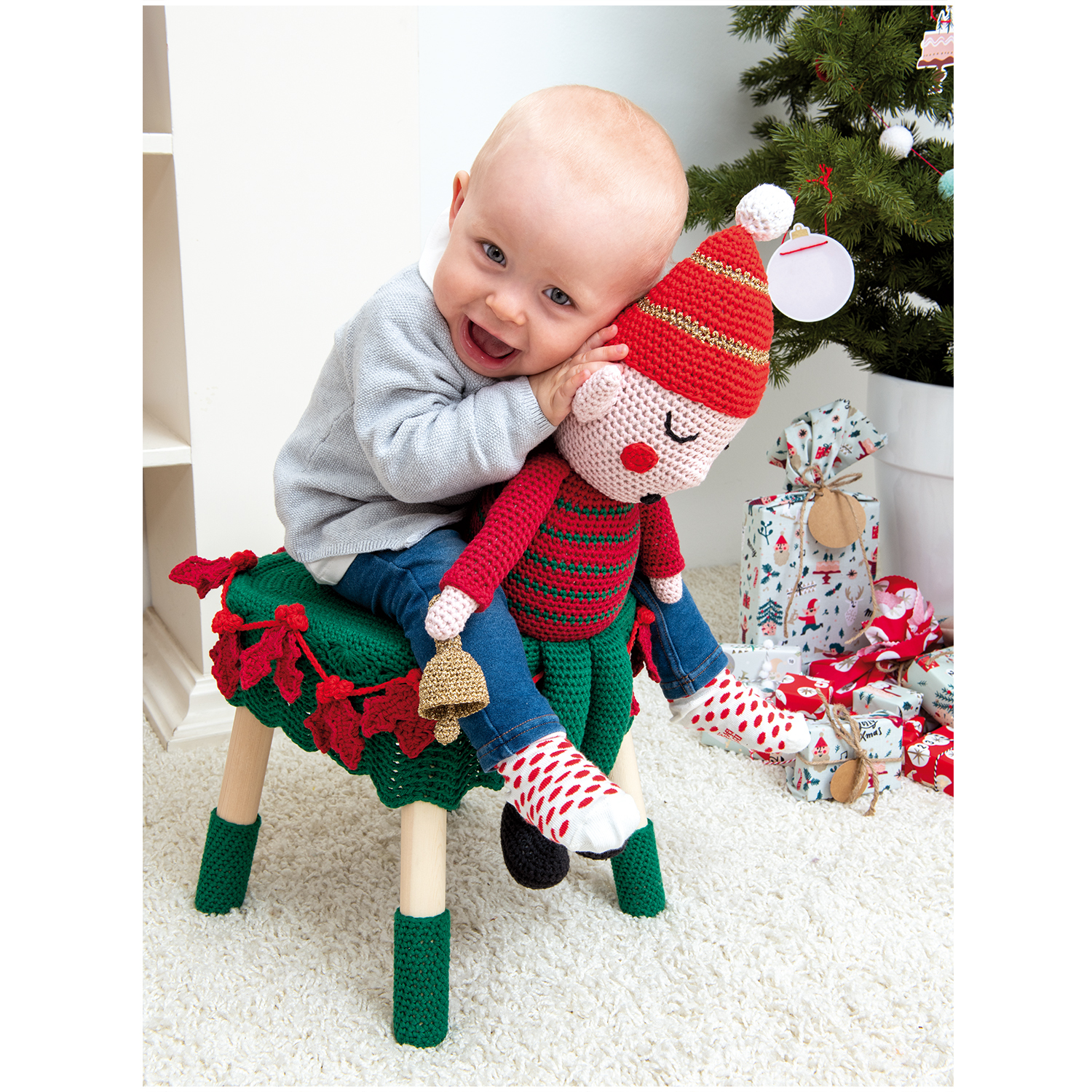 Creative Lamé: Ein Baby umarmt ein gestricktes Spielzeug auf einem dekorierten Hocker vor einem Weihnachtsbaum.