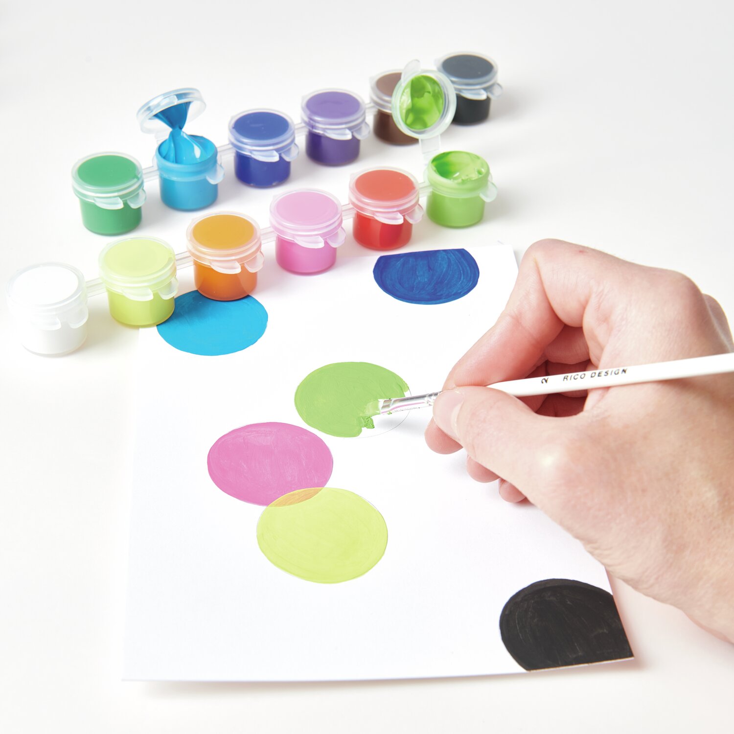 Acrylini Pods: Hand malt mit Pinsel bunte Kreise auf Papier, umgeben von 12x3,5ml Farbtöpfen.
