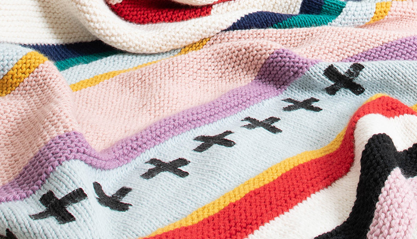 Strickanleitung Decke aus Essentials Mega Wool chunky: Bunte gestrickte Decke mit Streifen und Kreuzmustern in lebendigen Farben.