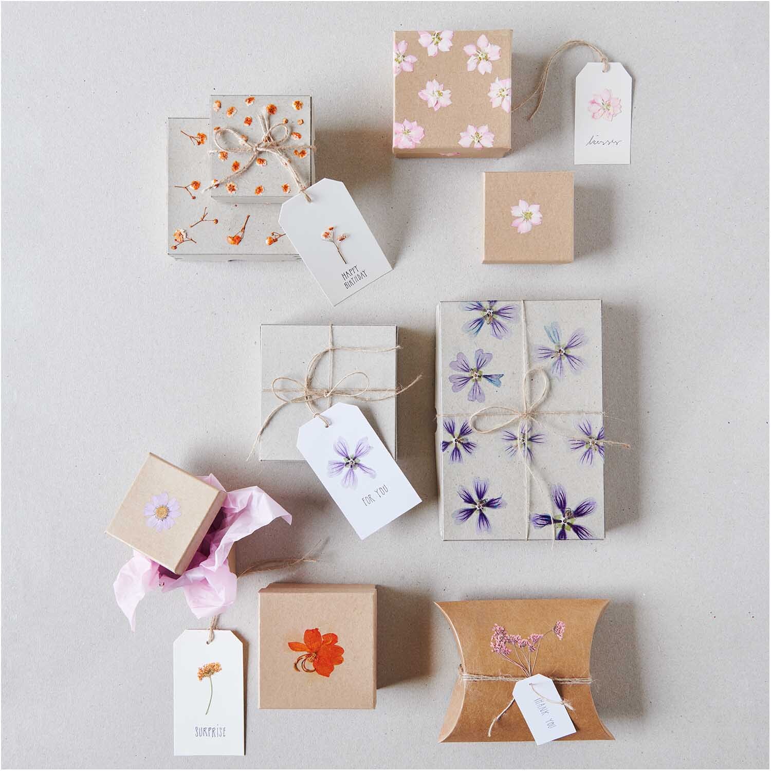 Quadratbox natur: Verschiedene Geschenkboxen mit floralen Mustern und Anhängern, arrangiert auf einer neutralen Oberfläche.