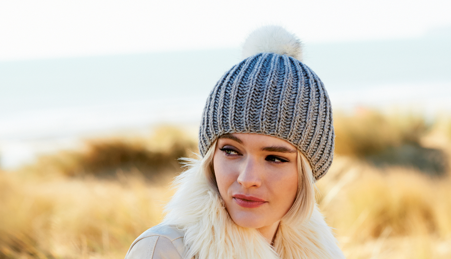 Strickanleitung Mütze mit Kunstfellpompon: Frau trägt graue Strickmütze mit weißem Pompon am Strand.