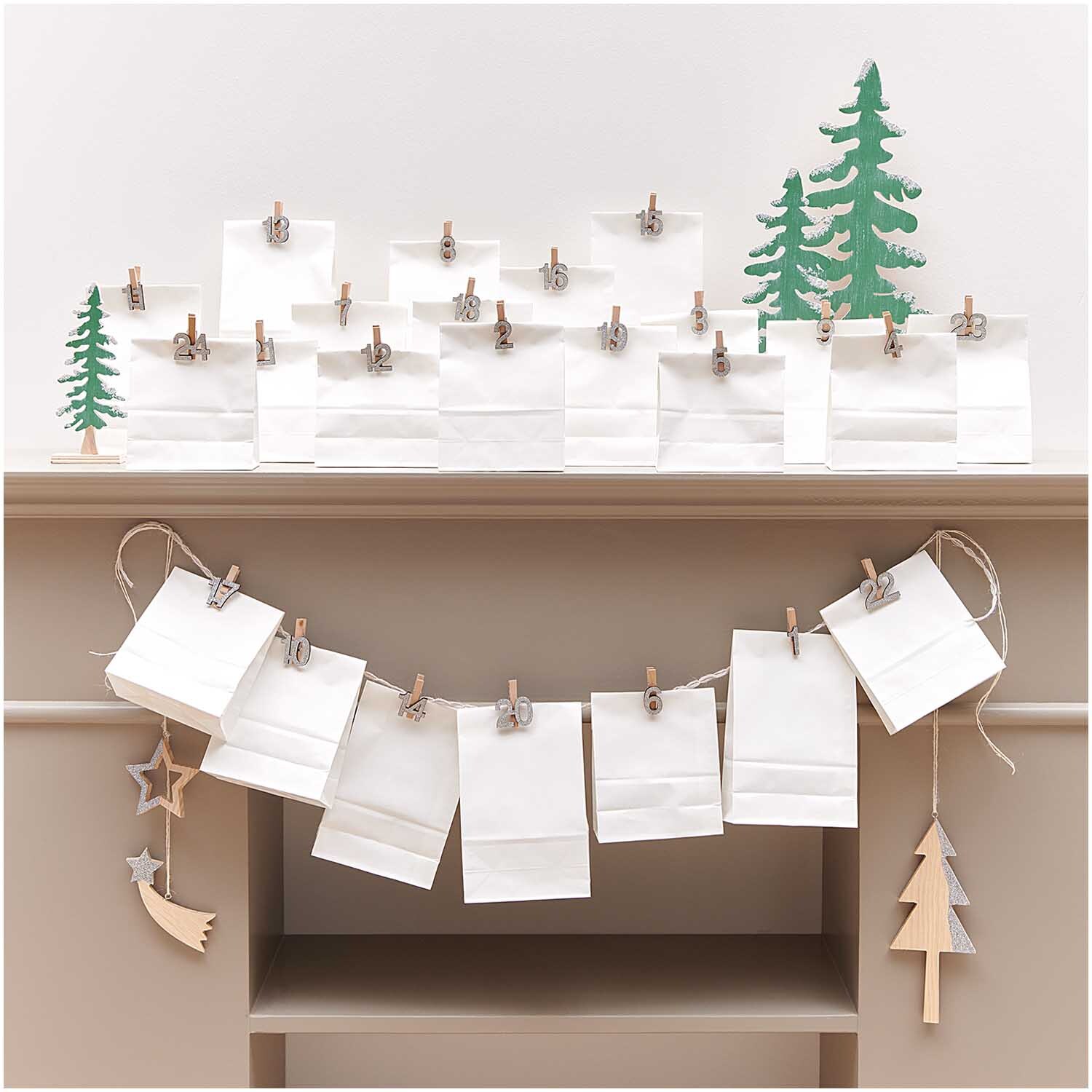 Holzclips Adventskalender Zahlen 1-24: Weiße Tüten mit goldenen und silbernen Glitzerzahlen, dekoriert auf einem Kaminsims mit Tannenbaum-Design.