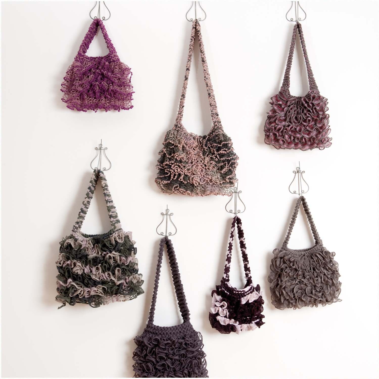 Strickanleitung Tasche aus Schalgarnen: Sieben handgefertigte Taschen in verschiedenen Designs und Farben, an Haken hängend.