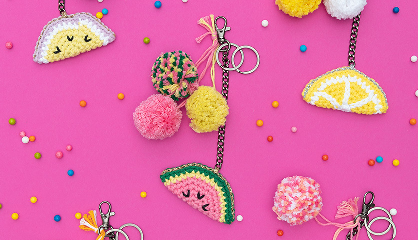 Häkelanleitung Obstanhänger: Bunte gehäkelte Fruchtanhänger und Pompons auf pinkem Hintergrund.