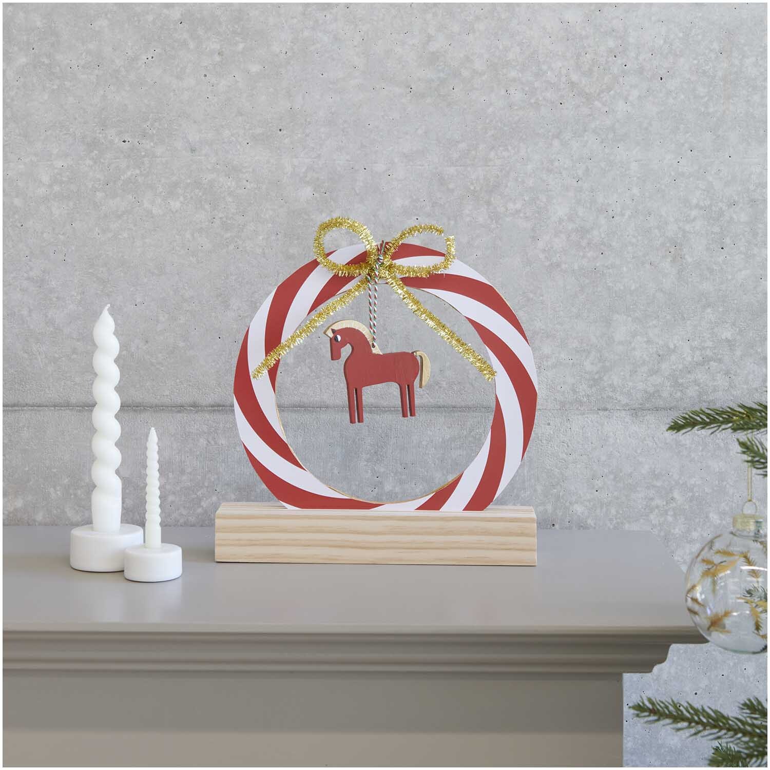 Paper Poetry Kartonring Candy Cane Ø 25cm mit rot-weißen Streifen und goldener Schleife, dekoriert mit einem Holzpferd, auf einem Holzsockel.