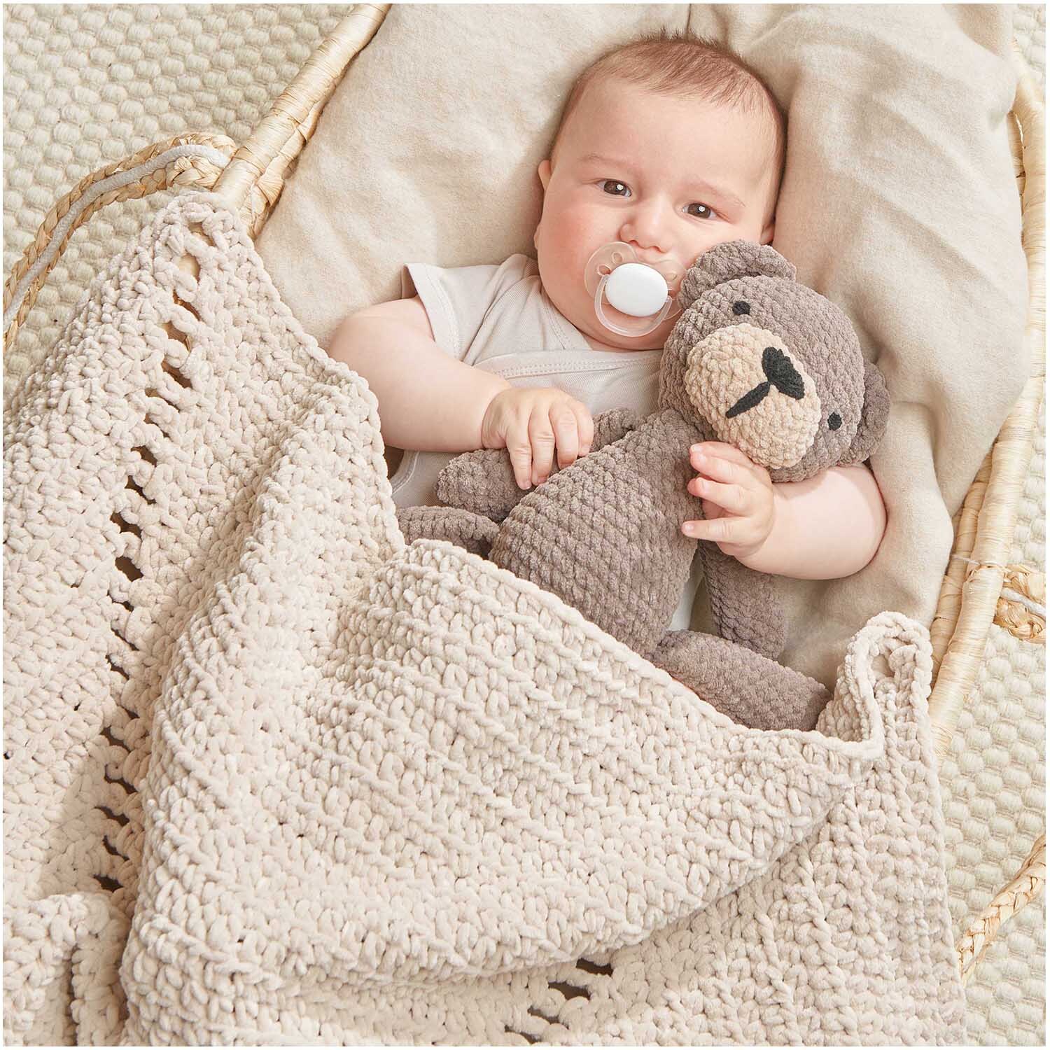 Creative Chenillove: Ein Baby liegt in einem Korb mit gehäkelter Decke und kuschelt mit einem Plüschtier.
