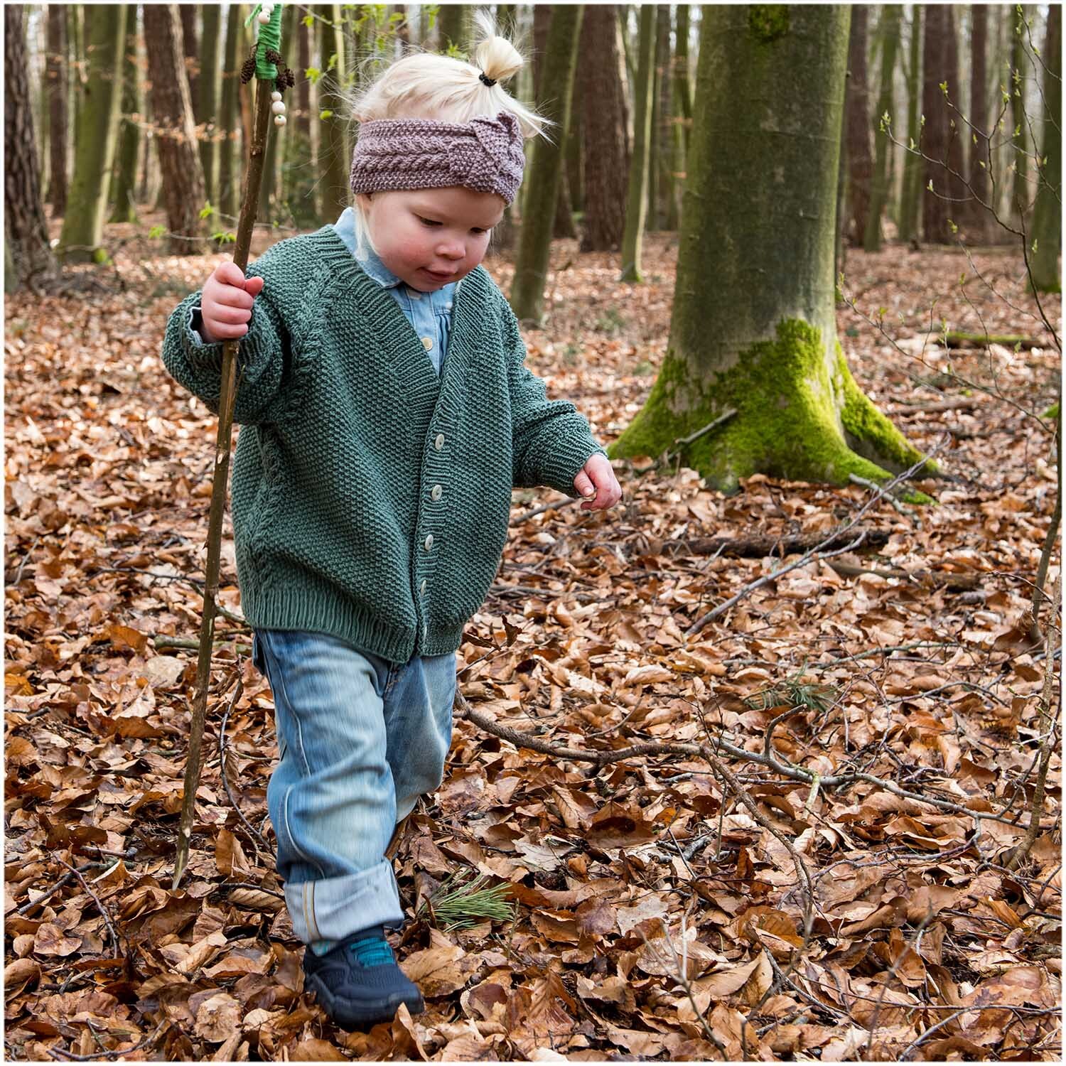 Essentials Merino dk: Kind im Wald mit Strickjacke in Efeu, trägt Jeans und Stirnband, Rico Design.