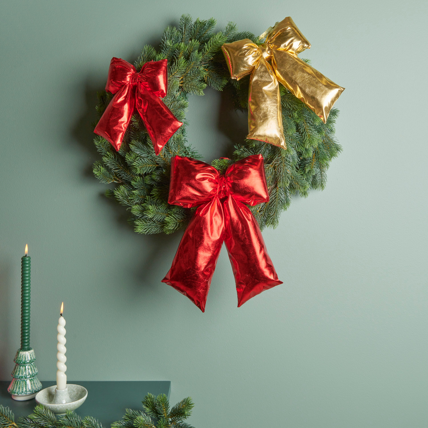 Weihnachtsschleife Rot: Festlicher Kranz mit roten und goldenen Schleifen, dekoriert mit grünen Kerzenhaltern und Spiralkerzen.