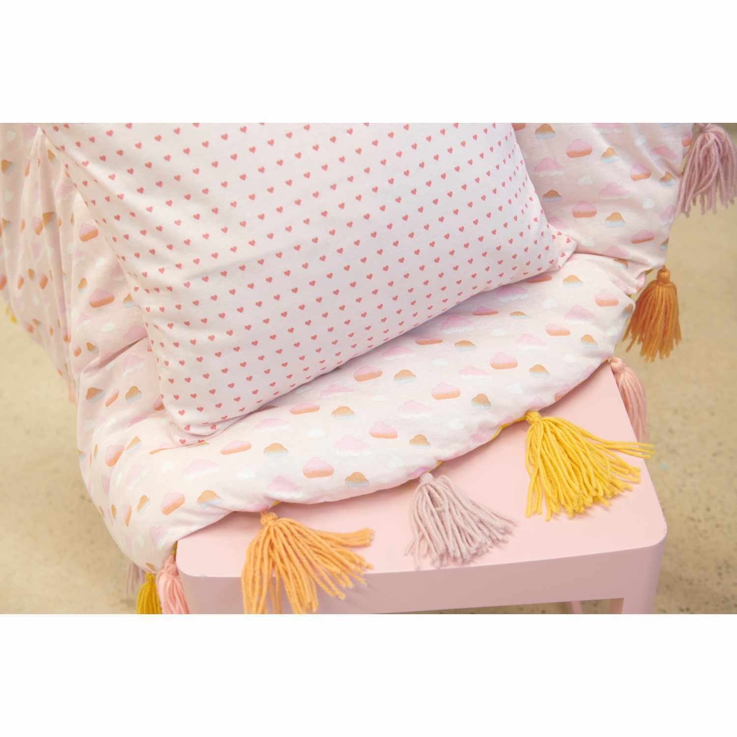 Jerseystoff Happy Baby Wolken rosa 145cm von Rico Design, zeigt rosa Wolkenmuster auf Stoff mit Kissen und Quasten.