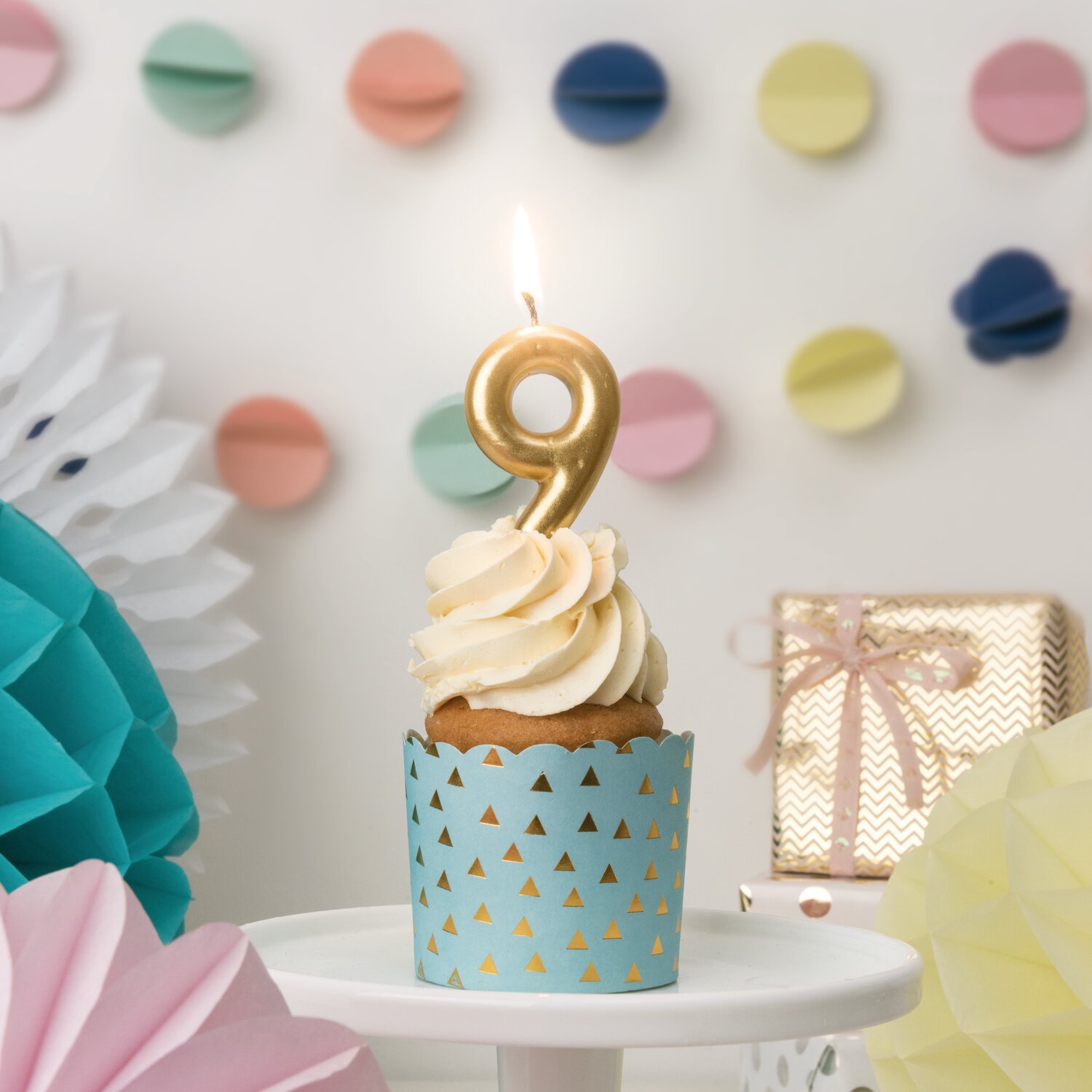 Zahlenkerze Gold von Rico Design auf Cupcake mit cremefarbenem Frosting, Partydeko mit bunten Wabenbällen und Geschenk im Hintergrund