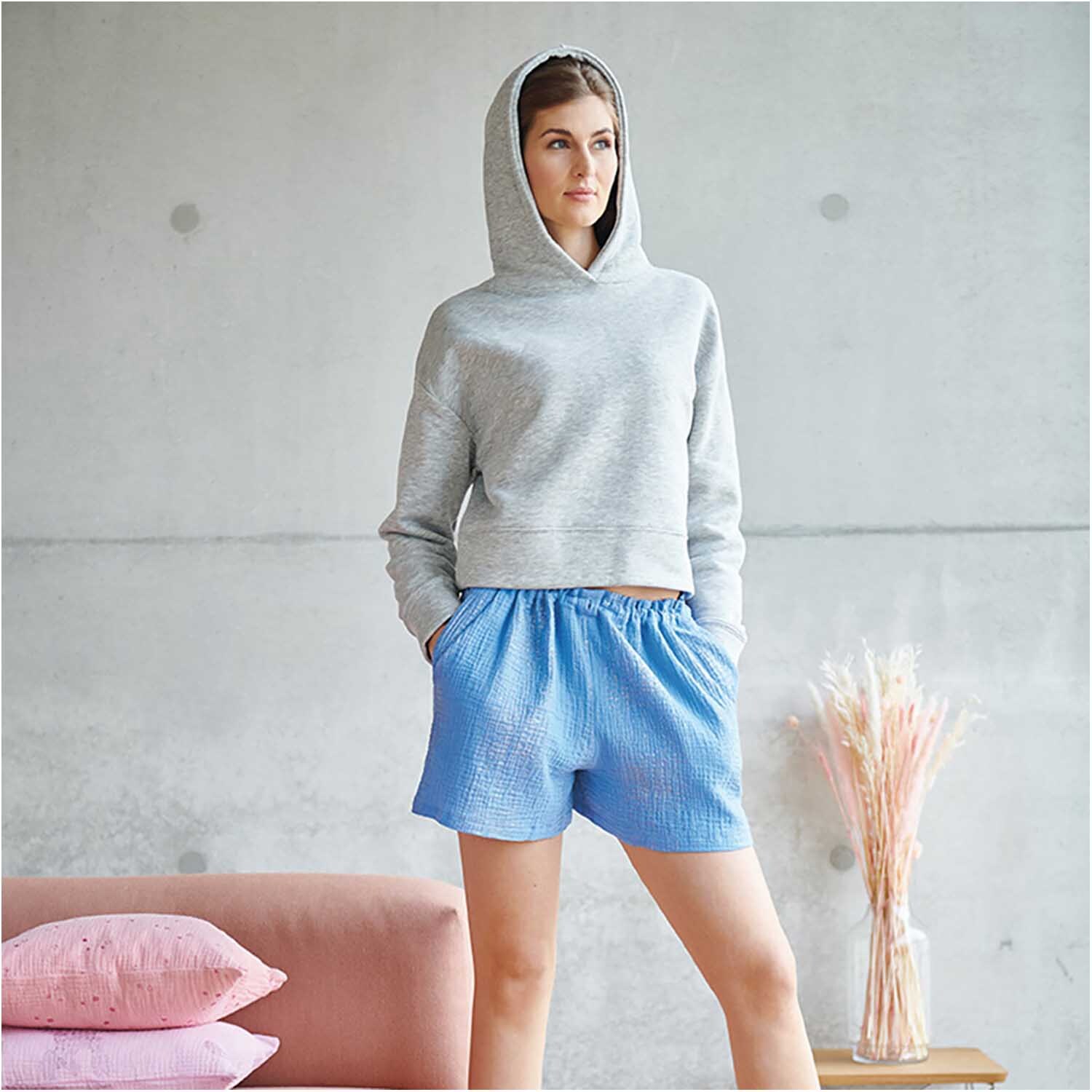 Stoffabschnitt Krinkelmusselin azur 50x130cm Hot Foil: Frau in grauem Hoodie und blauen Shorts aus Musselin, neben rosa Kissen und Trockenblumen.