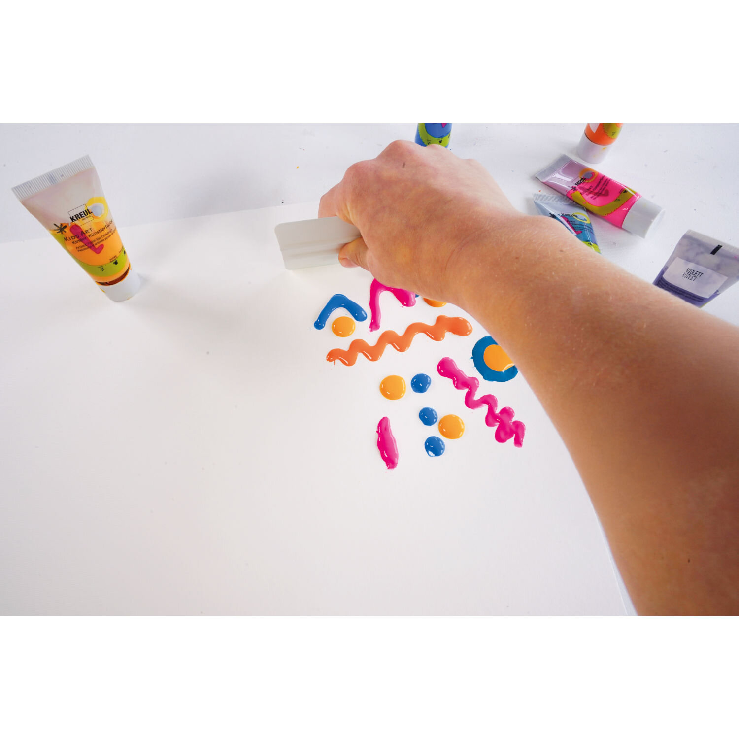 Squeegee Art Set von KREUL, Hand verteilt bunte Farbstreifen und Punkte mit Rakel auf weißem Papier, um kreative Wischbilder zu gestalten