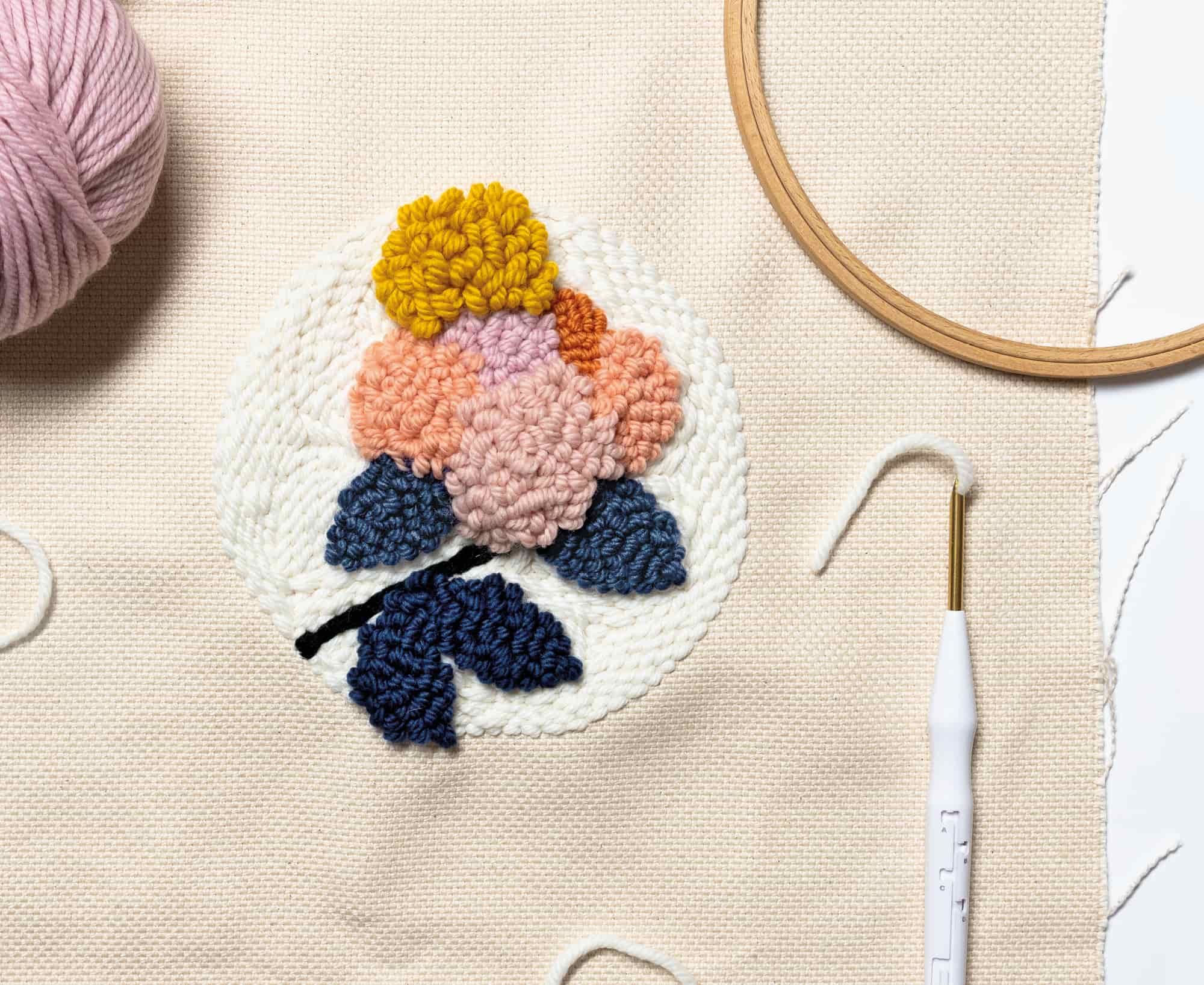Ein Blumenmuster ist mit der Punch Needle auf Stoff gestickt. Punch Needle, Stickrahmen und Wollknäuel liegen daneben. 