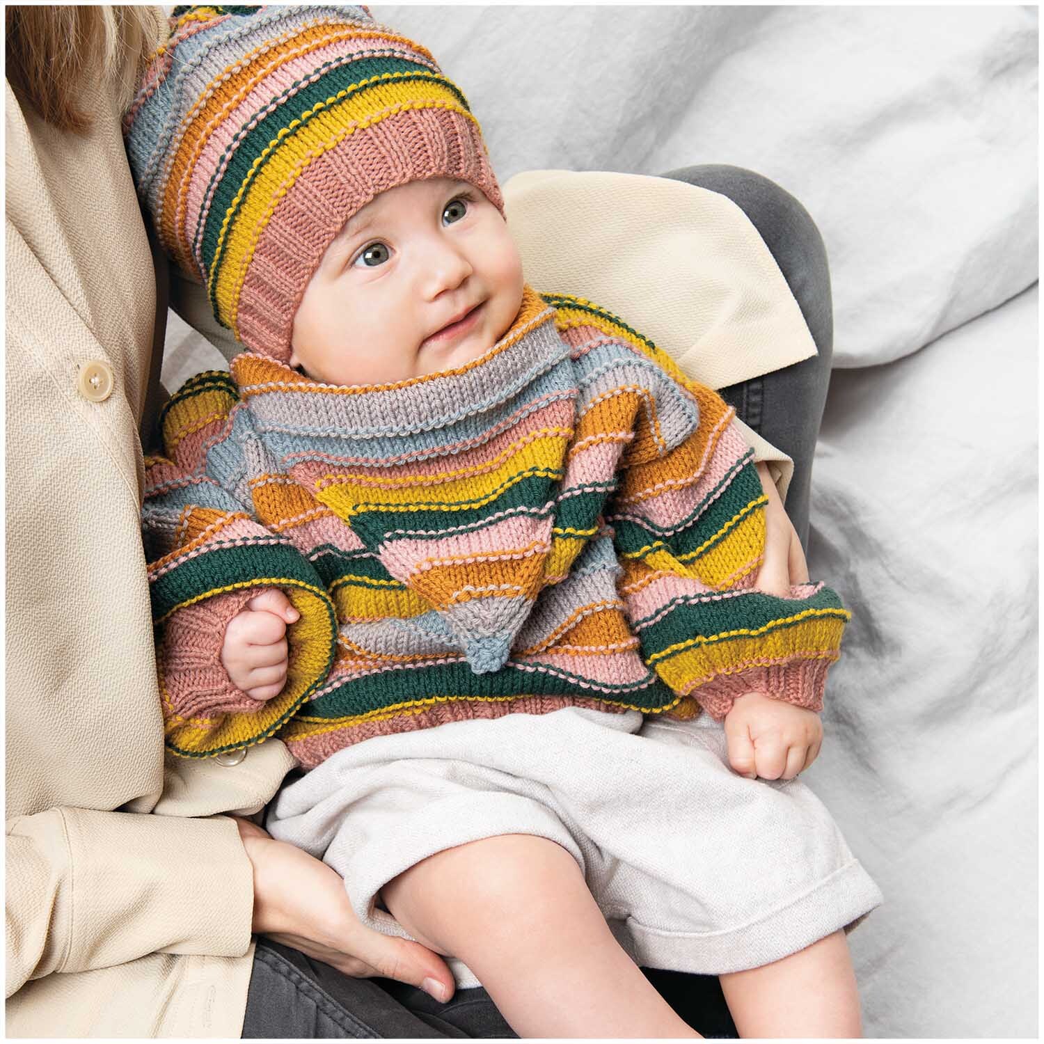 Rico Baby Classic dk: Ein Baby trägt einen gestreiften Pullover und Mütze in bunten Farben auf dem Schoß eines Erwachsenen.