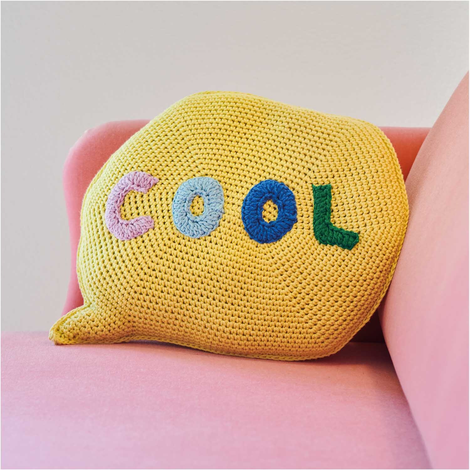 Creative Cotton aran Kissen auf rosa Sofa, gelb gehäkelt mit buntem "COOL"-Schriftzug.