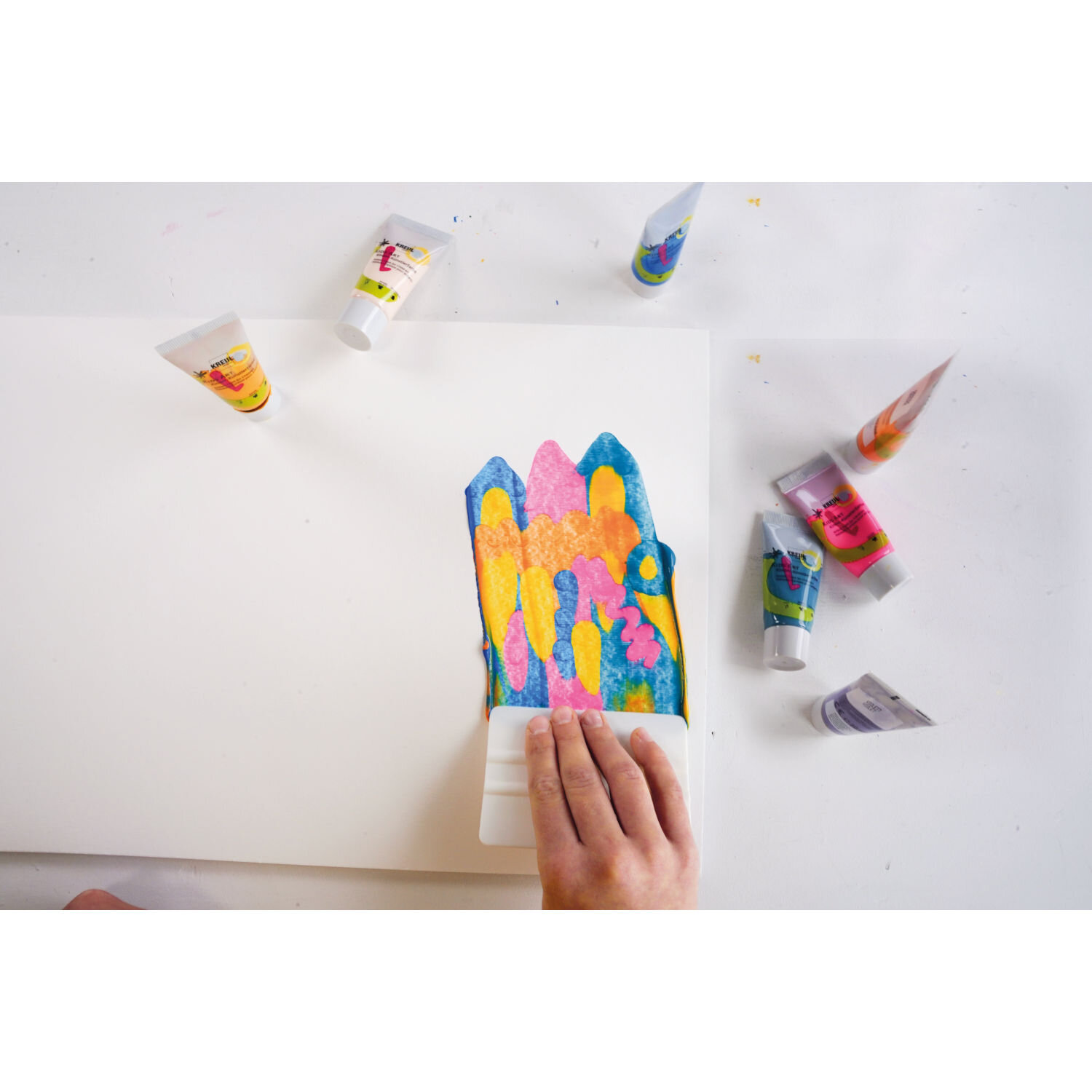 Squeegee Art Set von KREUL, Hand zieht Kunststoffrakel über bunt aufgetragene Acrylfarben auf weißem Papier, daneben mehrere Farbtuben verteilt