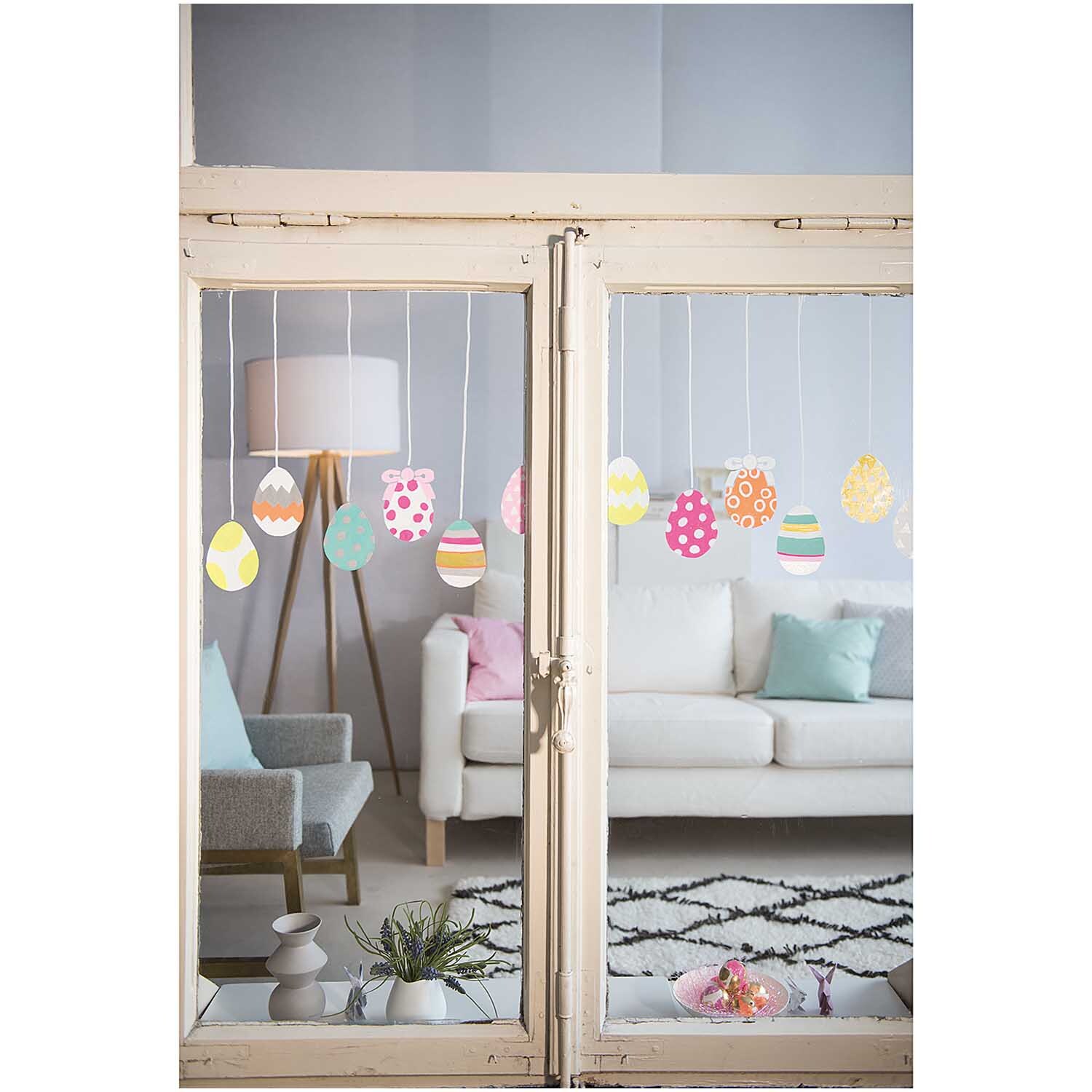 DIY Set Fenster gestalten Mermaid von Rico Design: Bunte Fensterdekoration mit hängenden Eiern in einem gemütlichen Wohnzimmer.