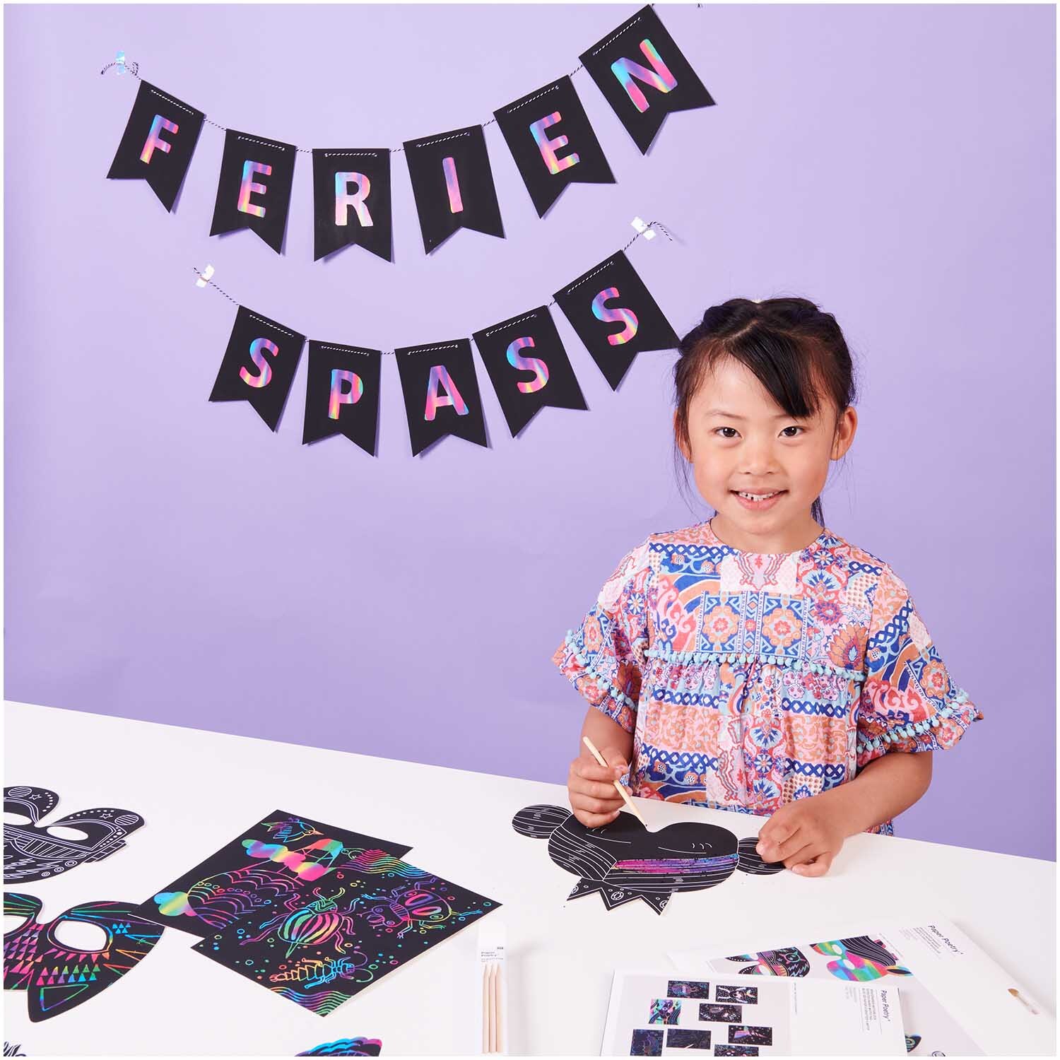 Paper Poetry Kratzpapier Girlande mit 12 Wimpeln und Holzkratzer, dekoriert in bunten Farben. Ein Kind bastelt fröhlich am Tisch.