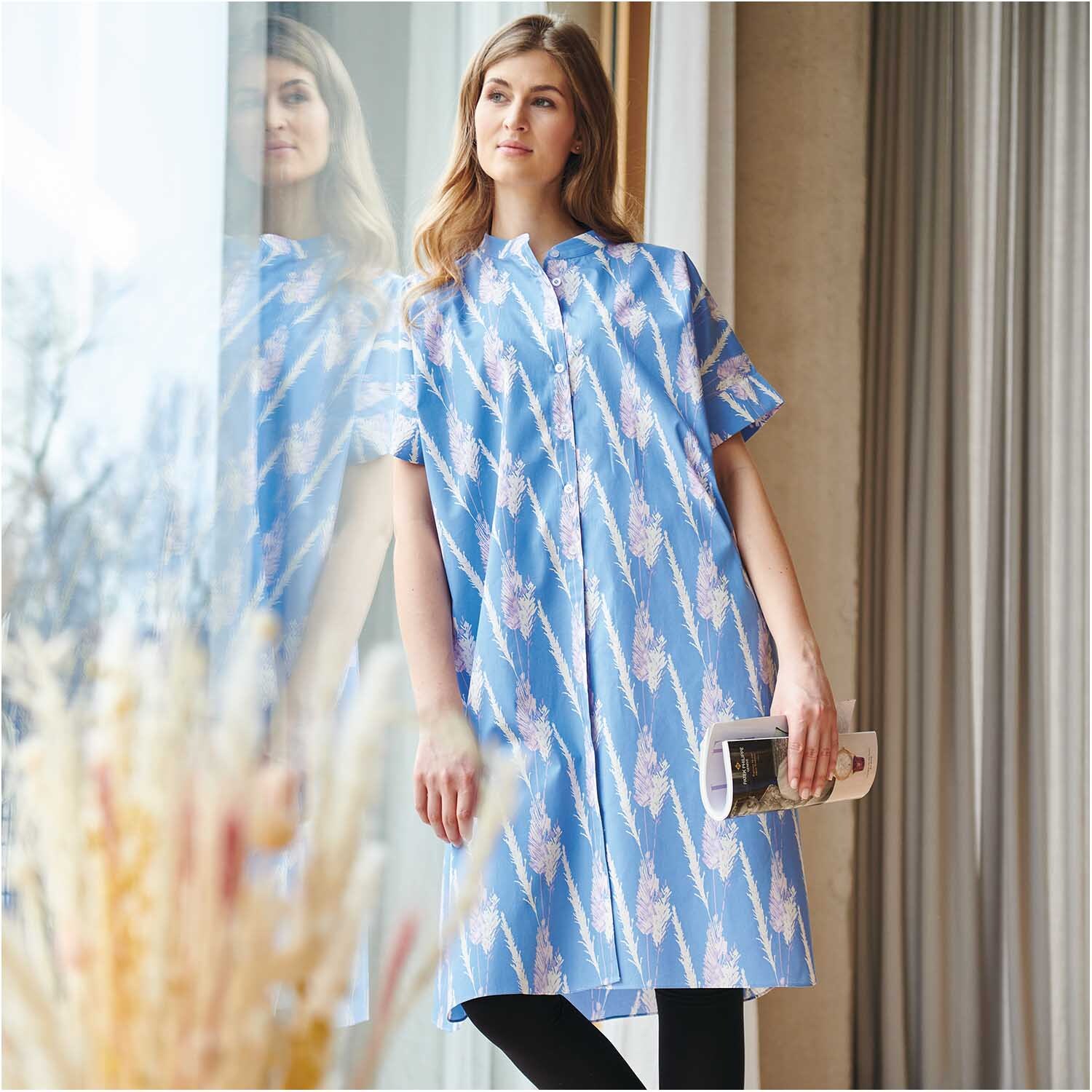 Meterware Druckstoff Pampasgras azur: Frau in azurblauem Kleid mit Pampasgras-Muster, steht am Fenster.