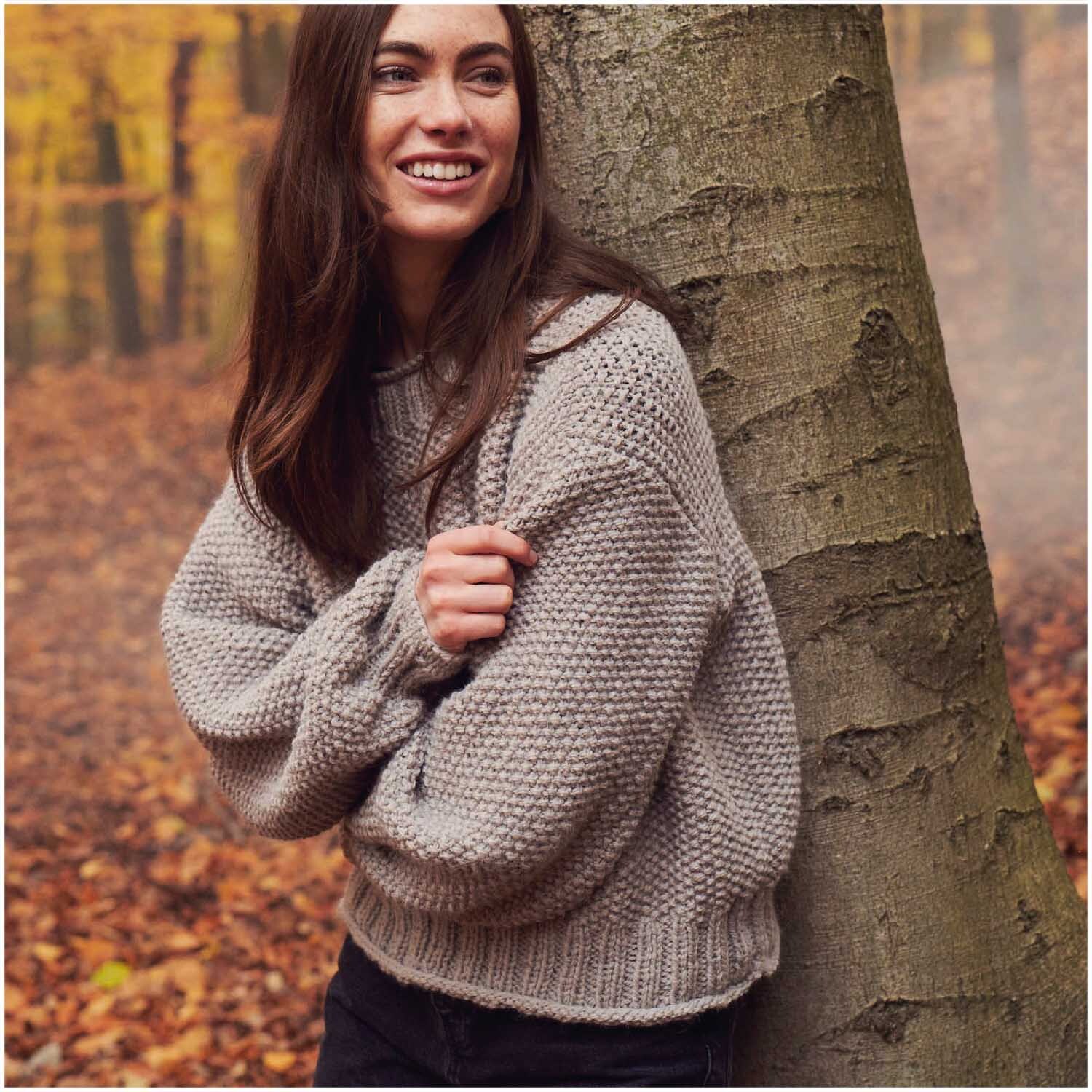 Essentials Alpaca Twist Chunky: Frau in grauem Strickpullover lehnt lächelnd an Baum im herbstlichen Wald.