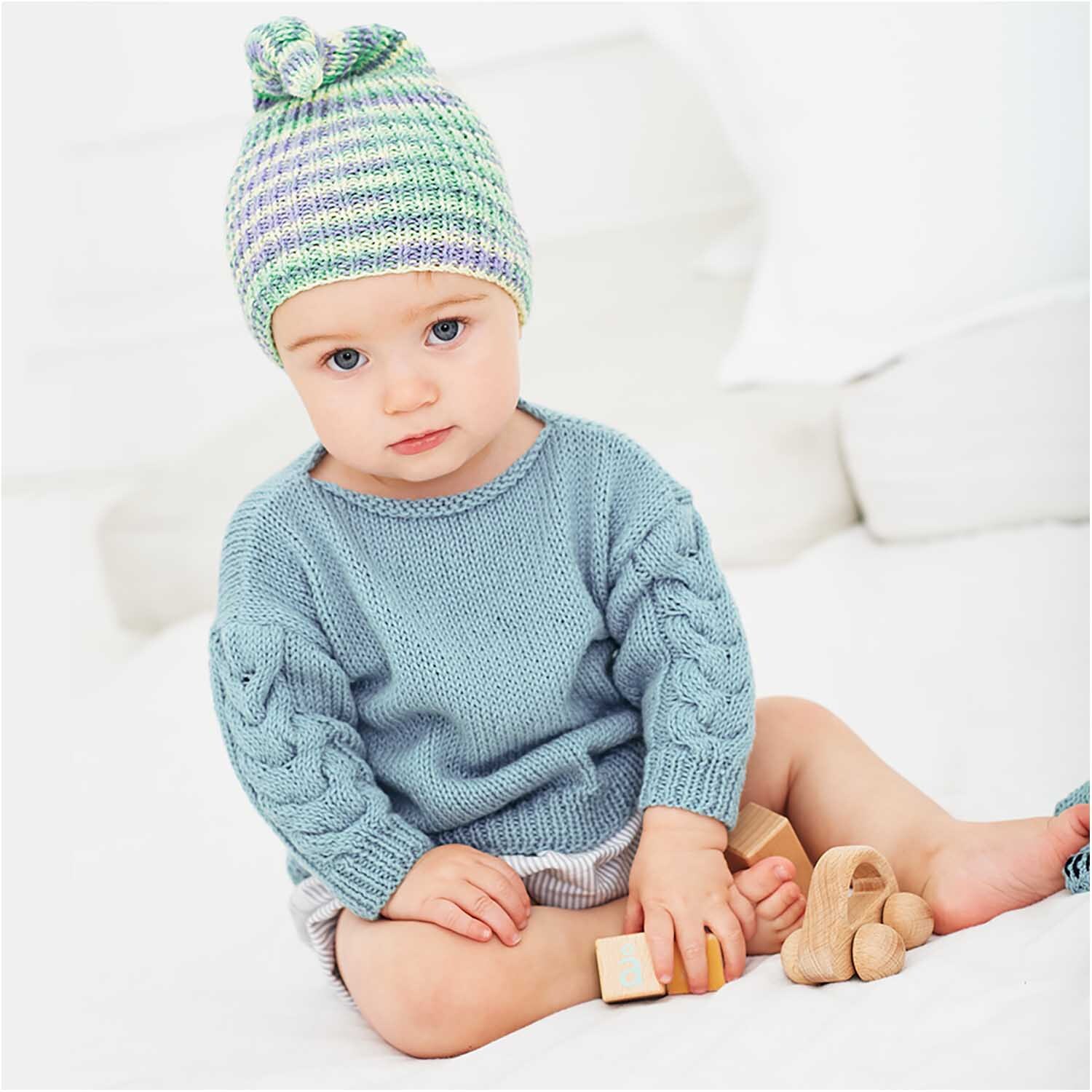 Rico Baby Cotton Soft Print dk: Kleinkind in gestricktem Pullover und Mütze in Veilchen-Grün, spielt mit Holzspielzeug.