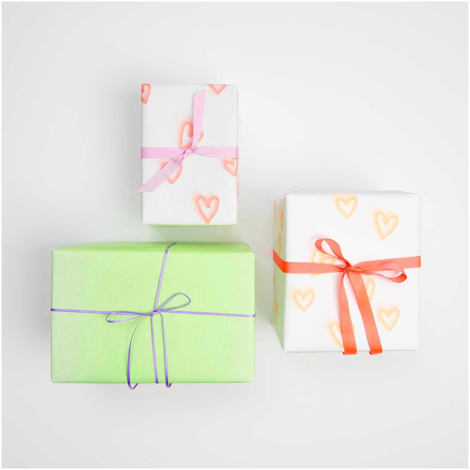 Paper Poetry Geschenkpapier zeigt weiße Pakete mit neonorangen Herzen und farbigen Schleifen.