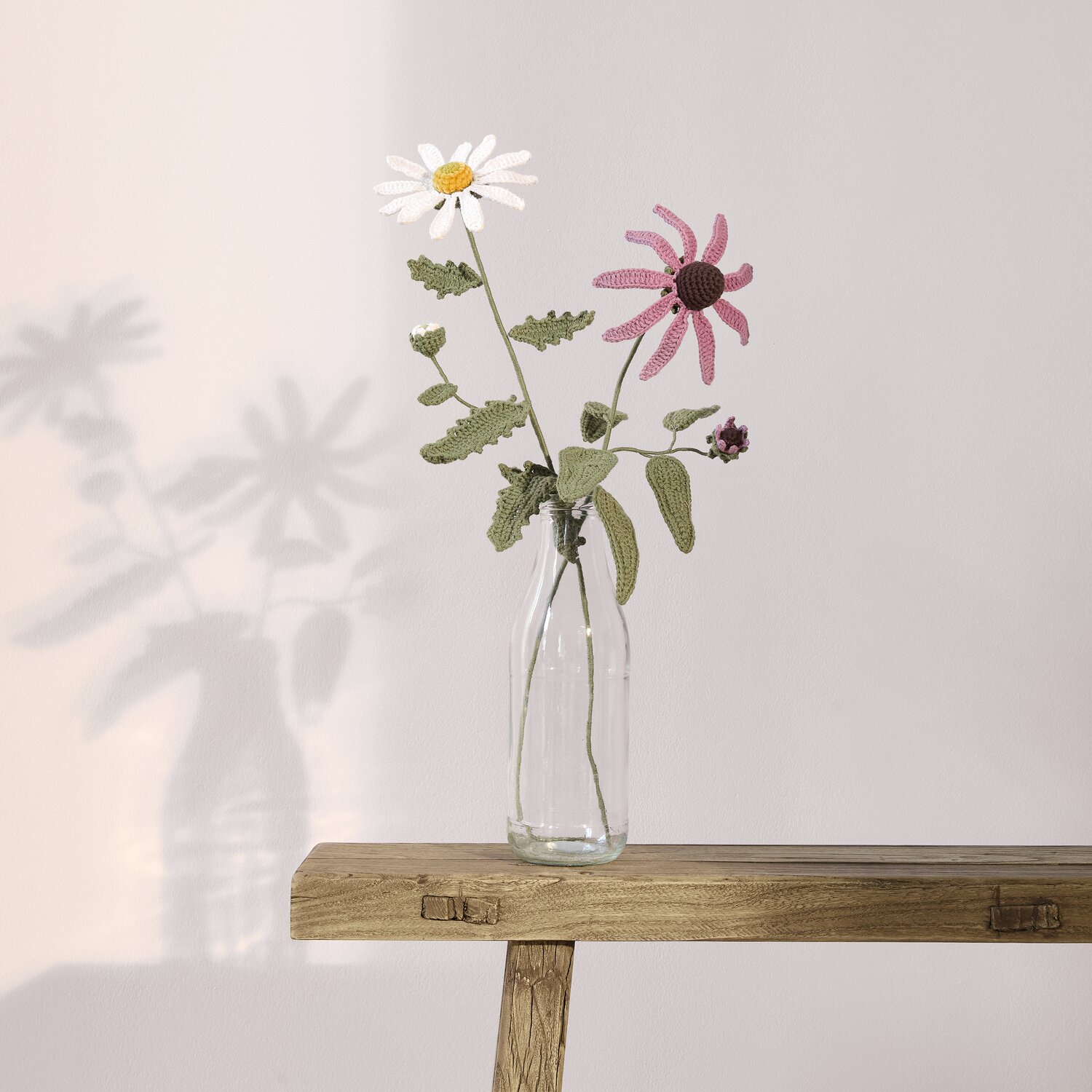 Ricorumi In the Meadow, gehäkelte Wiesenblumen in Glasvase auf Holzbänkchen, zarte Weiß- und Mauvetöne mit olivgrünen Blättern, dekorative Handarbeit