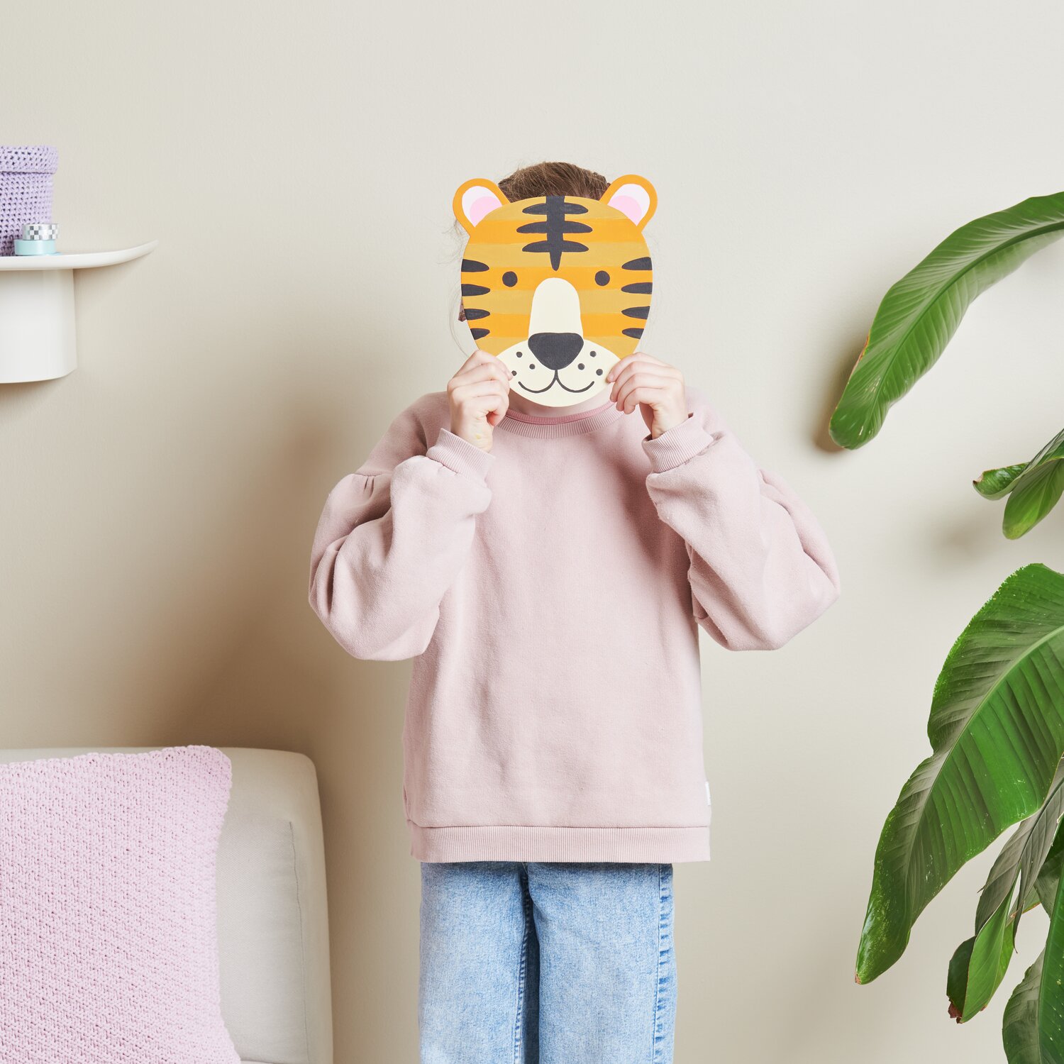 Malen nach Zahlen Tiger: Kind hält eine Tiger-Maske vor dem Gesicht, trägt rosa Pullover und Jeans.