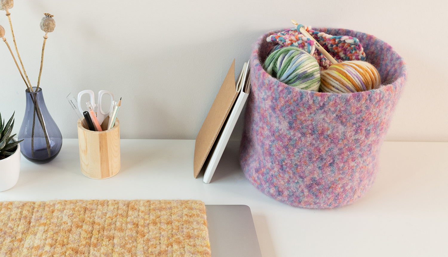 Strick- und Häkelanleitung für einen Filz-Korb, gefüllt mit buntem Garn, auf einem Schreibtisch neben Notizbüchern.