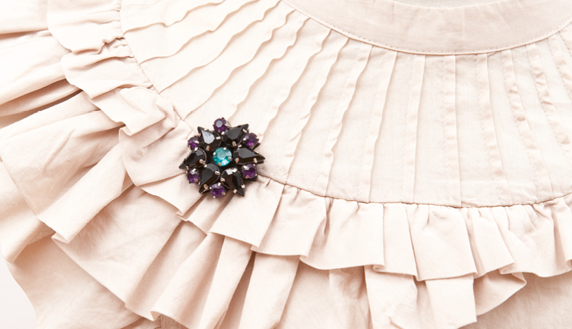 Anleitung Dekorative Brosche basteln: Eine elegante Brosche mit violetten und blauen Steinen auf einem beigen, gefalteten Stoff.