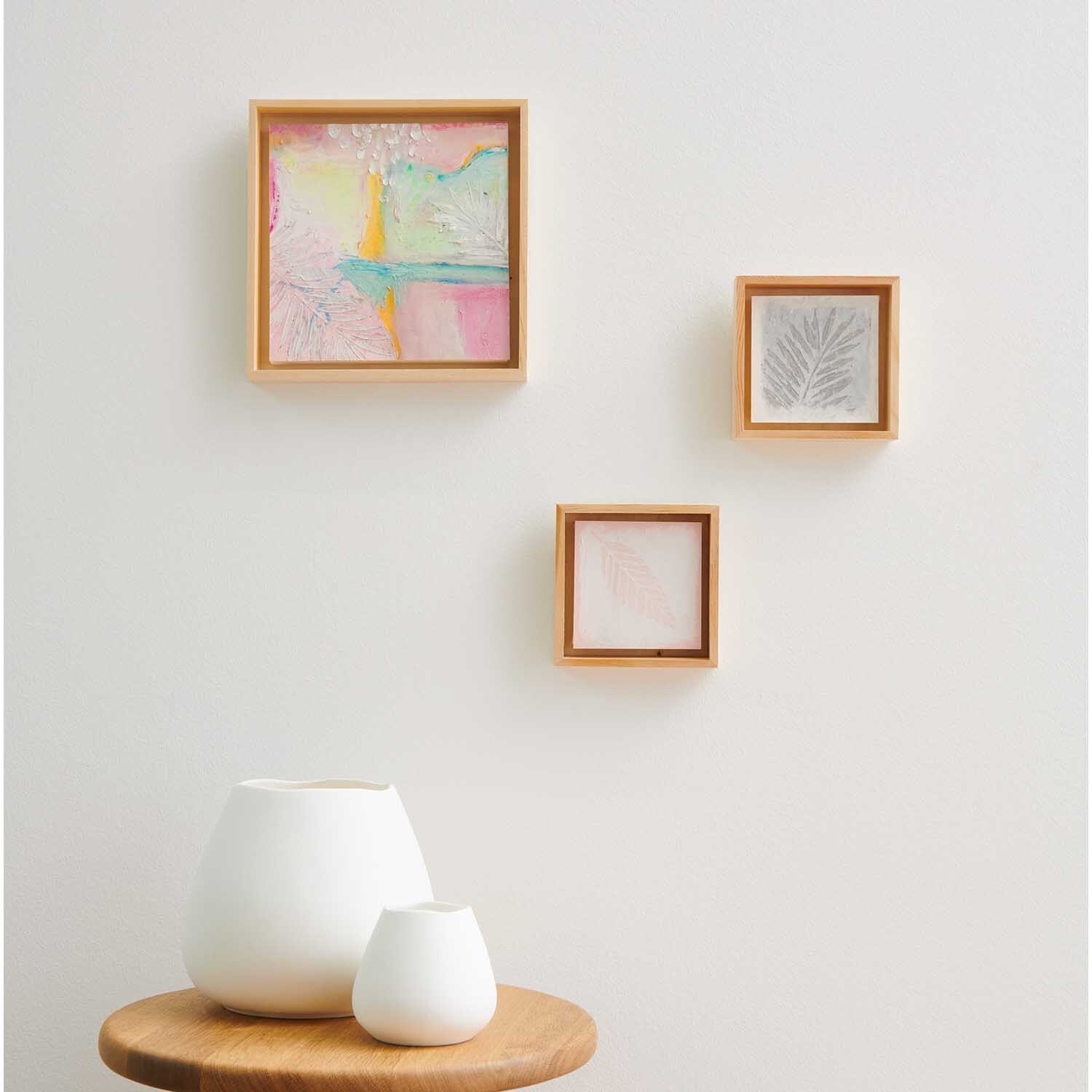 Ölpastellkreide Pastell & Neon 12 Farben von Rico Design: Drei gerahmte Kunstwerke an einer weißen Wand über zwei weißen Vasen auf einem Holztisch.