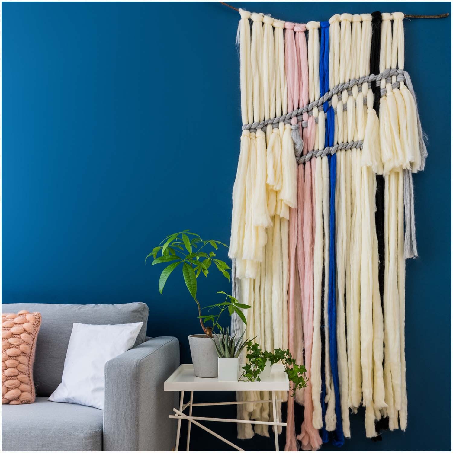 Creative Cocon Wandbehang in Creme, Gelb, Aqua, Blau, Rosa und Grau vor blauer Wand. Dekorativ neben grauem Sofa und Pflanzen.