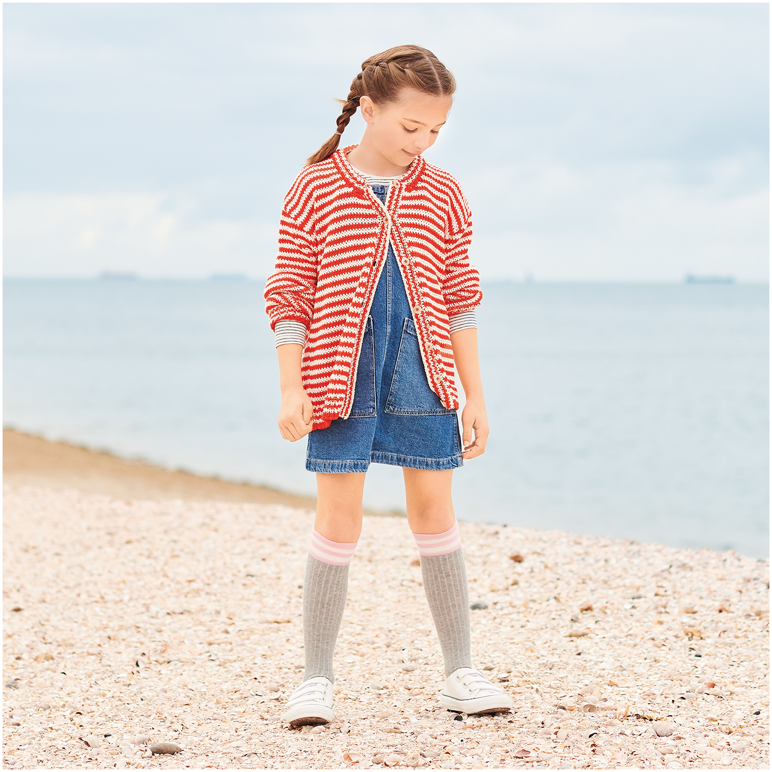 Essentials Organic Cotton aran: Mädchen am Strand in gestreifter Strickjacke und Jeanskleid, Erdbeerrot.