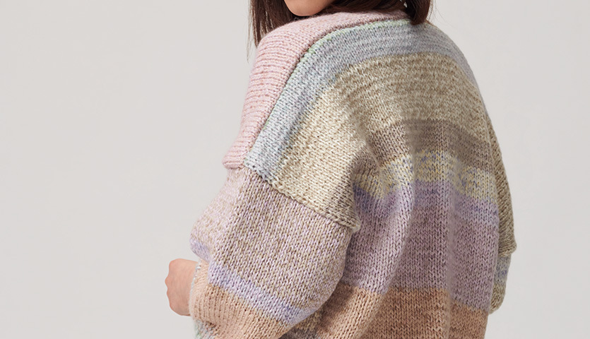 Strickanleitung Jacke aus Creative Chic-Unique: Frau trägt pastellfarbene, gestreifte Strickjacke. Rückenansicht zeigt weiche Textur und sanfte Farbübergänge.