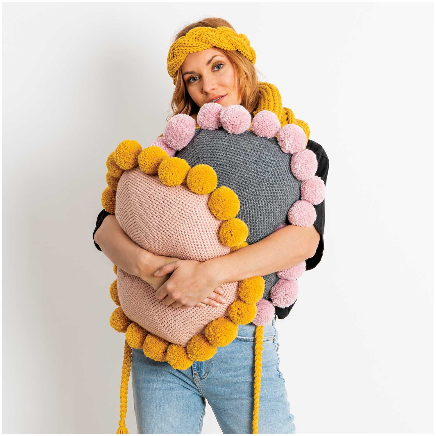 Essentials Mega Wool chunky: Frau hält gestricktes Kissen mit bunten Pompons, trägt Stirnband.