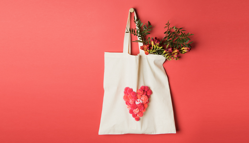 Bastelanleitung Baumwolltasche mit Pompons: Eine cremefarbene Tasche mit roten Pompons in Herzform, gefüllt mit Blumen, auf rotem Hintergrund.