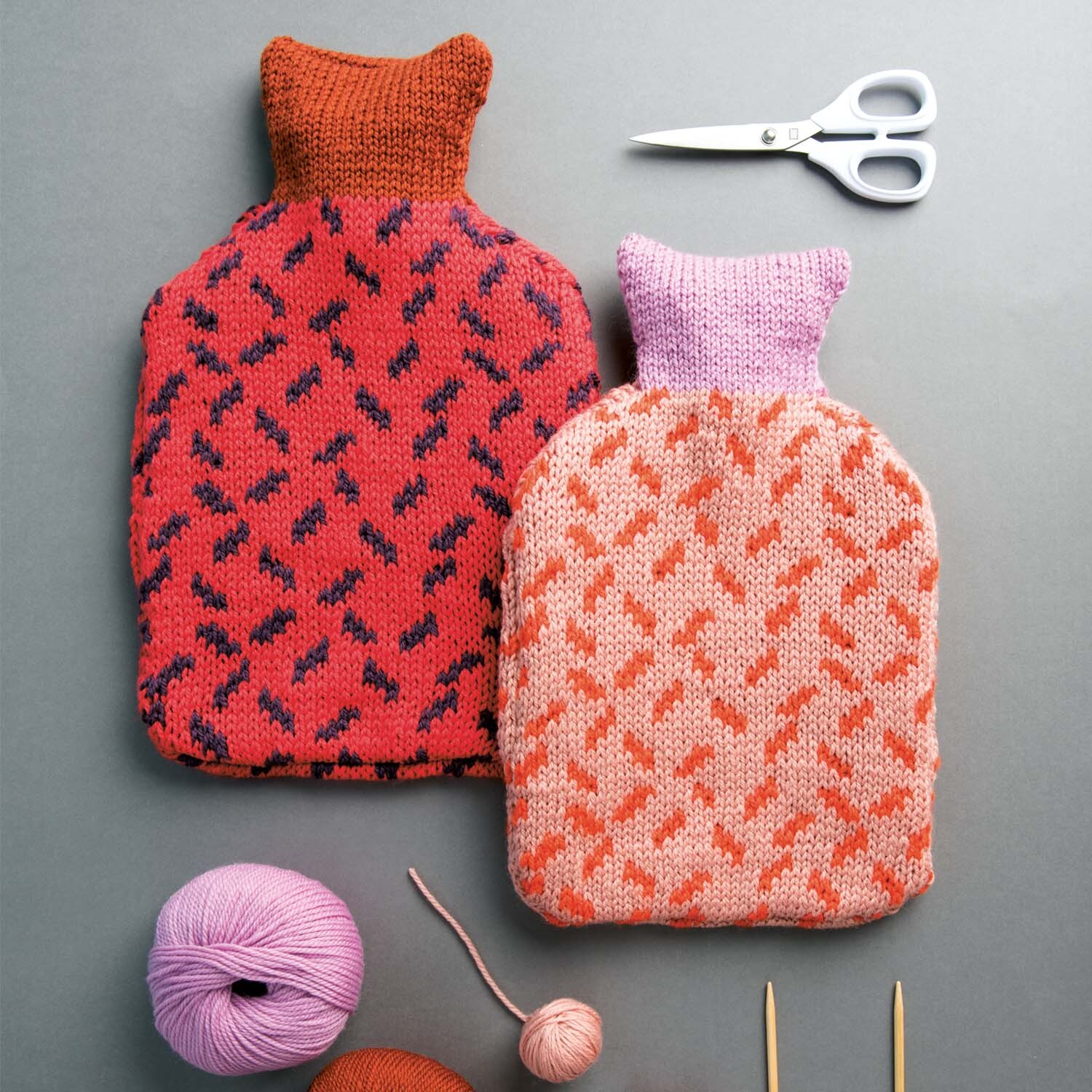 Strickanleitung Wärmflaschenhülle: Zwei gestrickte Wärmflaschenhüllen in Rot und Rosa mit Strickzubehör.