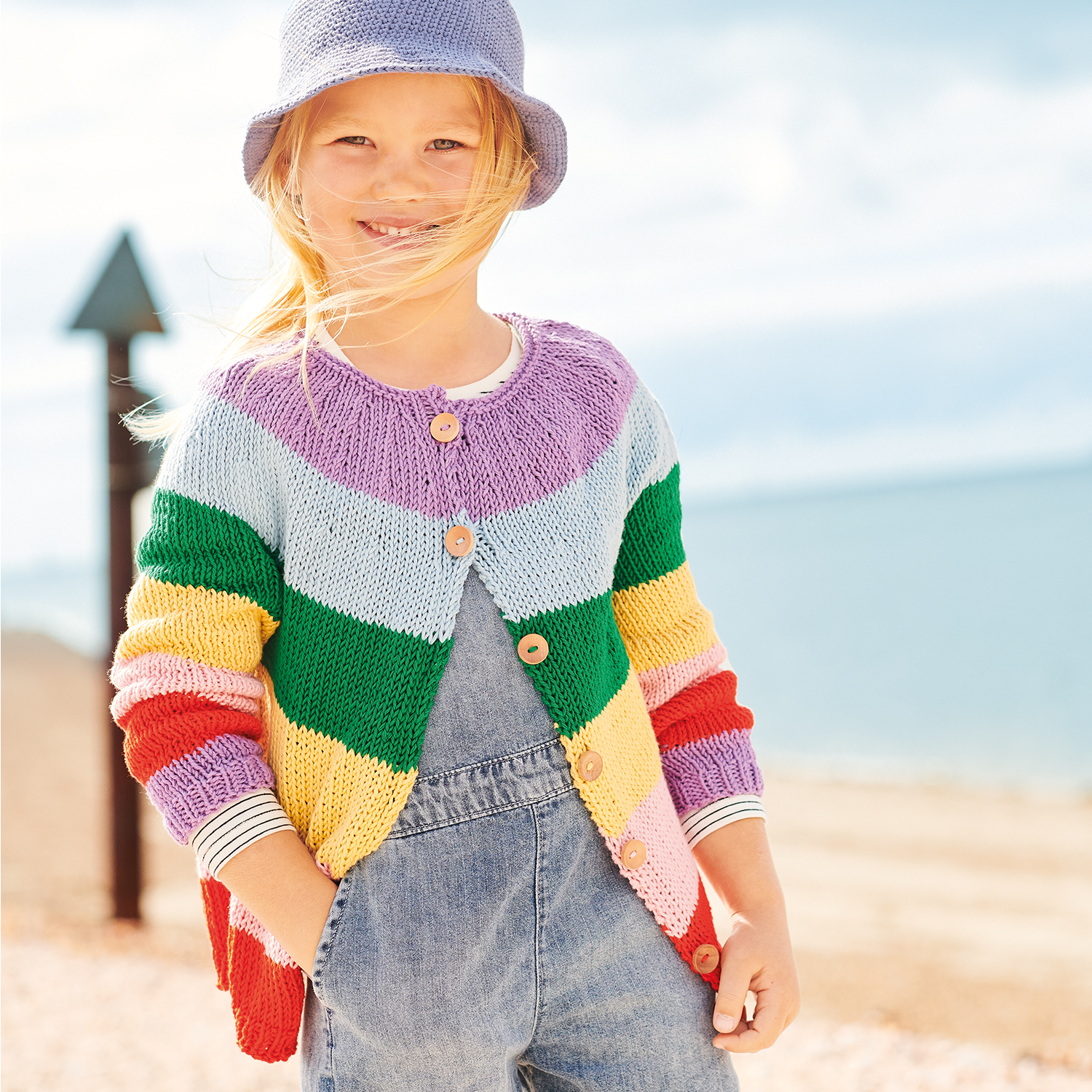 Strickanleitung Jacke aus Essentials Organic Cotton aran: Kind trägt bunte Strickjacke am Strand.