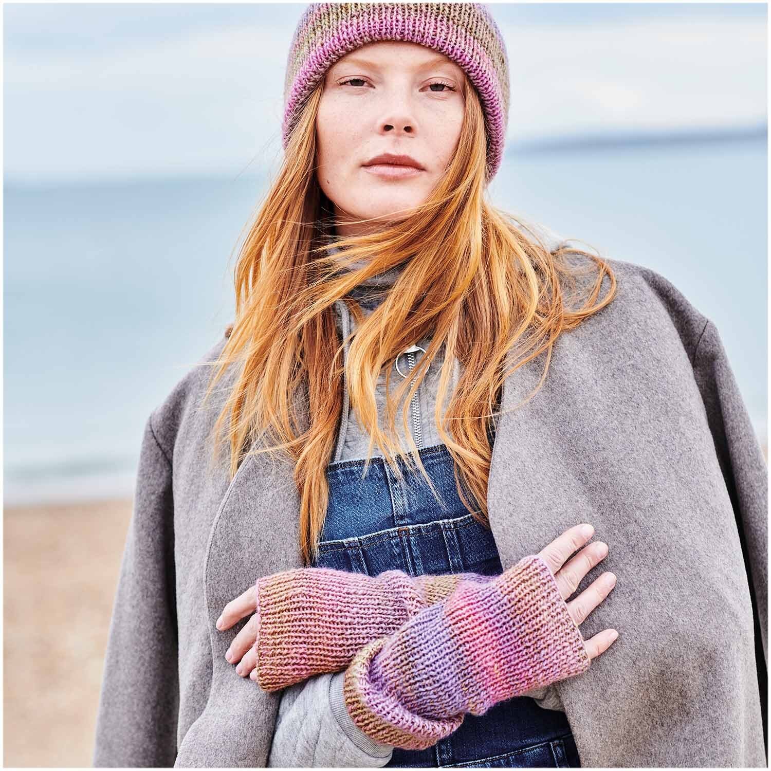 Creative Melange aran Wonderball: Frau am Strand trägt gestrickte Mütze und Handschuhe in Flieder-Pink von Rico Design.