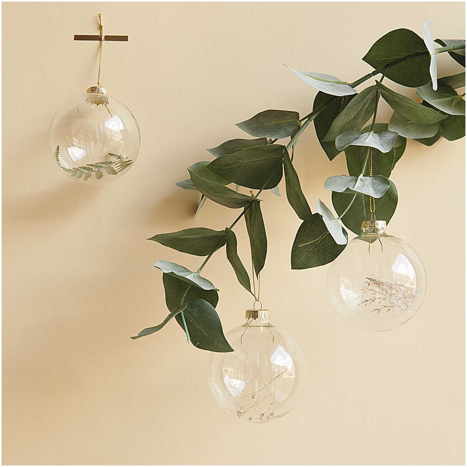 Glaskugel Baumschmuck mit Trockenfarn von Rico Design, hängend an einem Zweig mit grünen Blättern.