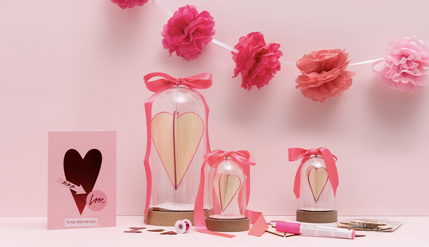 Bastelanleitung Dekohaube mit Steckherzen: Drei Glasglocken mit Herzen, rosa Schleifen und Dekoration auf rosa Hintergrund.