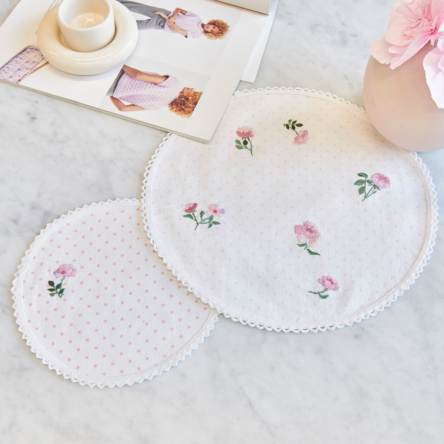 Stick and Stitch Rosen, 15 Stück: Zwei bestickte, runde Stoffuntersetzer mit Rosenmotiv auf weißem Hintergrund, dekorativ auf Marmorplatte arrangiert.