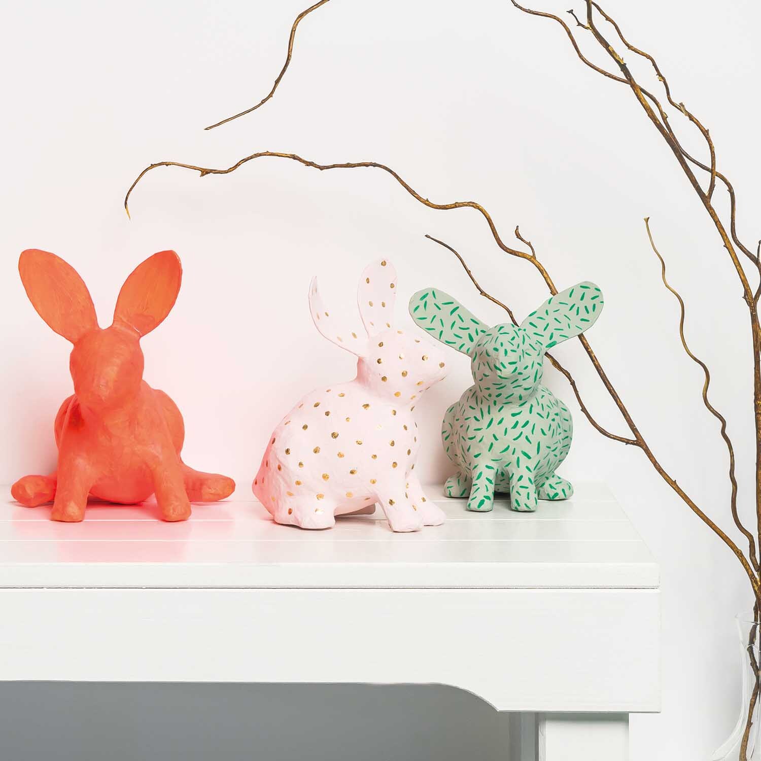 Anleitung Pappmaschee-Hasen mit Acrylfarbe gestalten: Drei bunte Hasenfiguren auf einem Tisch, dekoriert mit Zweigen im Hintergrund.