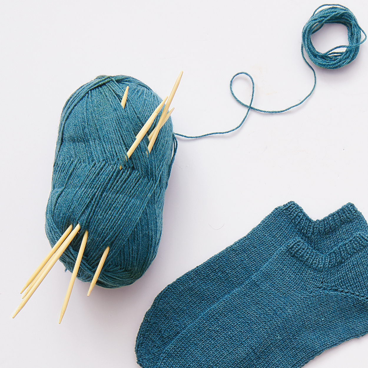Ein Paar gestrickte Sneakersocken aus einem blauen Garn, welches daneben liegt mit mehreren Holzstricknadeln. Flatlay von Produkten.