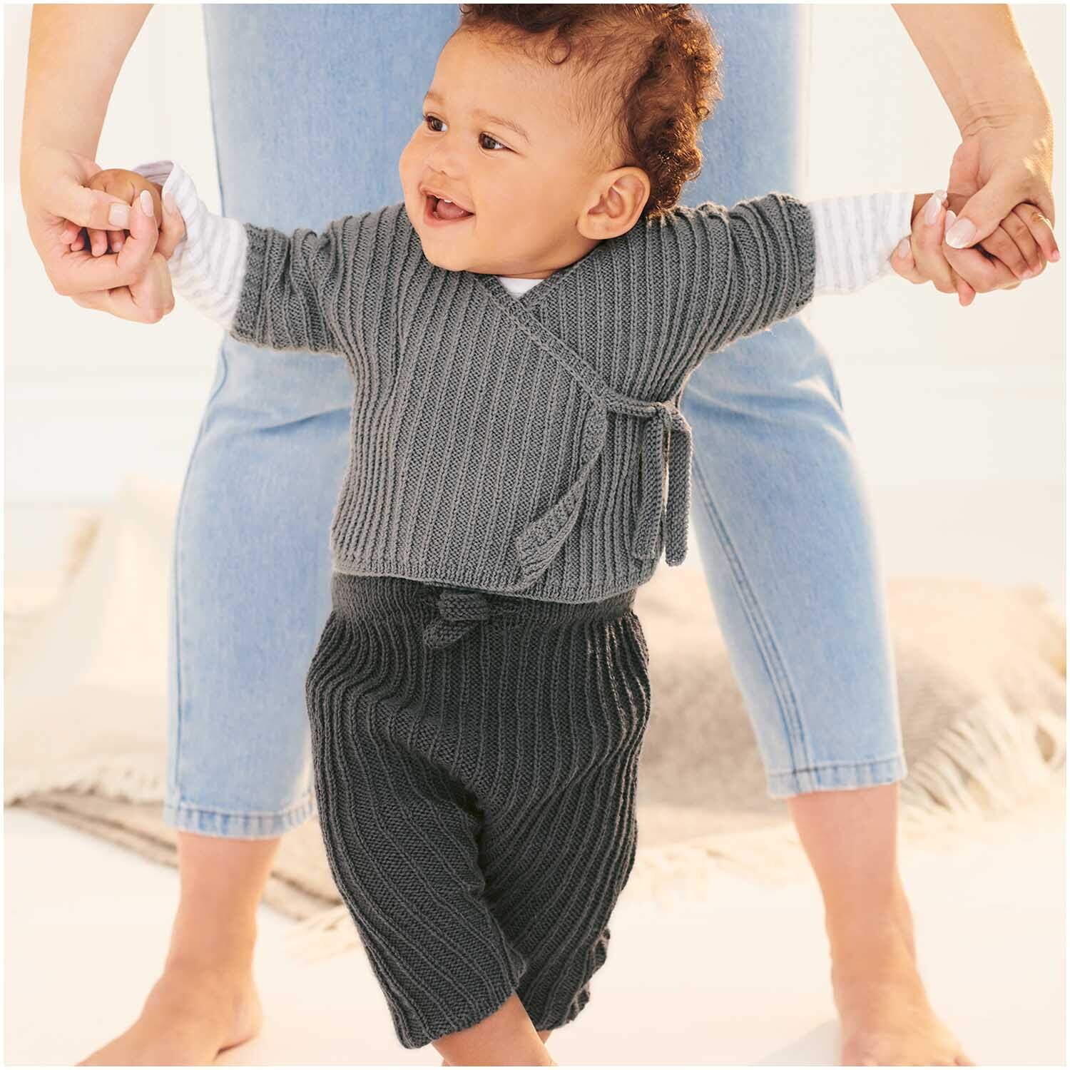 Baby Merino: Ein lächelndes Baby in grauer Strickkleidung von Rico Design, gehalten von einer erwachsenen Person.