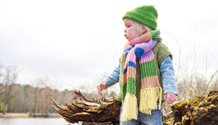 Strickanleitung Schal aus Essentials Soft Merino aran: Kind mit buntem Schal und grünem Hut steht draußen auf einem Baumstamm.