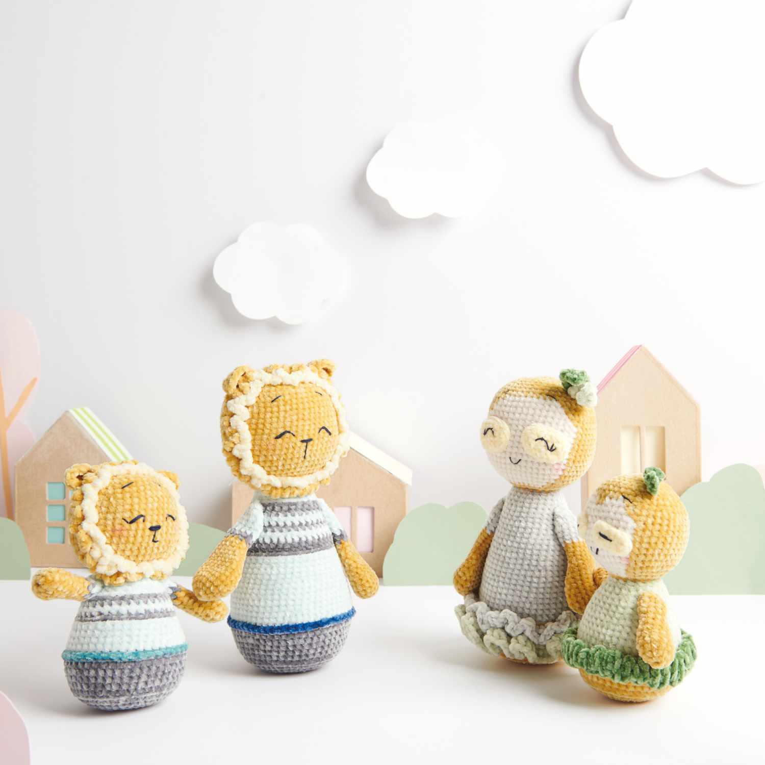 Häkelset Papa Löwe und Linus aus Ricorumi Mini Me: Zwei gehäkelte Löwenfiguren in Pastelltönen vor einem Hintergrund mit Wolken und Häusern.