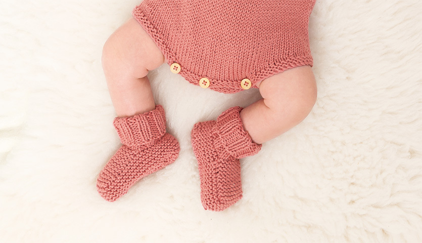 Strickanleitung Babysocken aus Baby Cotton Soft dk: Baby trägt rosa gestrickte Socken und passendes Outfit auf weißem Fell.