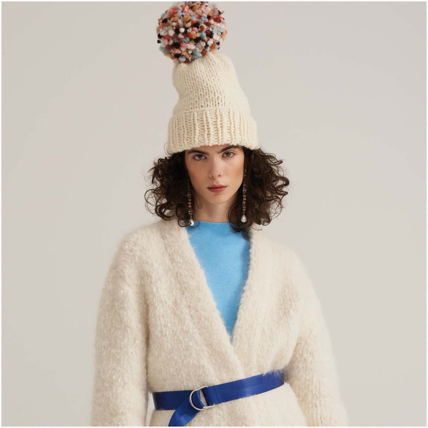 Fashion Fine Fur Super Chunky: Frau in cremefarbenem Strickmantel mit blauem Gürtel und bunter Bommelmütze.